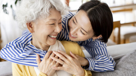 Two healthy Japanese women are smiling and hugging each other.They are sitting on the sofa in their living room.
