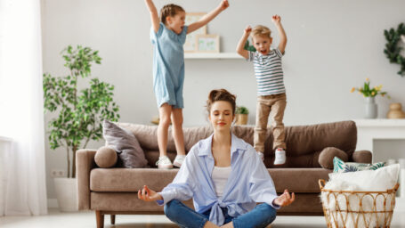 Happy mother with closed eyes meditating in lotus pose on floor trying to save inner harmony while excited children jumping on sofa and screaming in light spacious living room