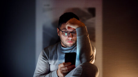 A 35-year-old guy is sitting in the dark room and using his smartphone.